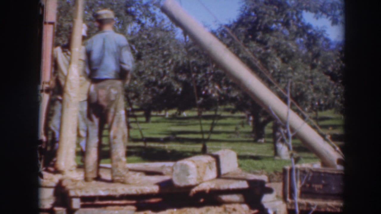Two men stand beside a portable saw rig and a large log in an orchard.