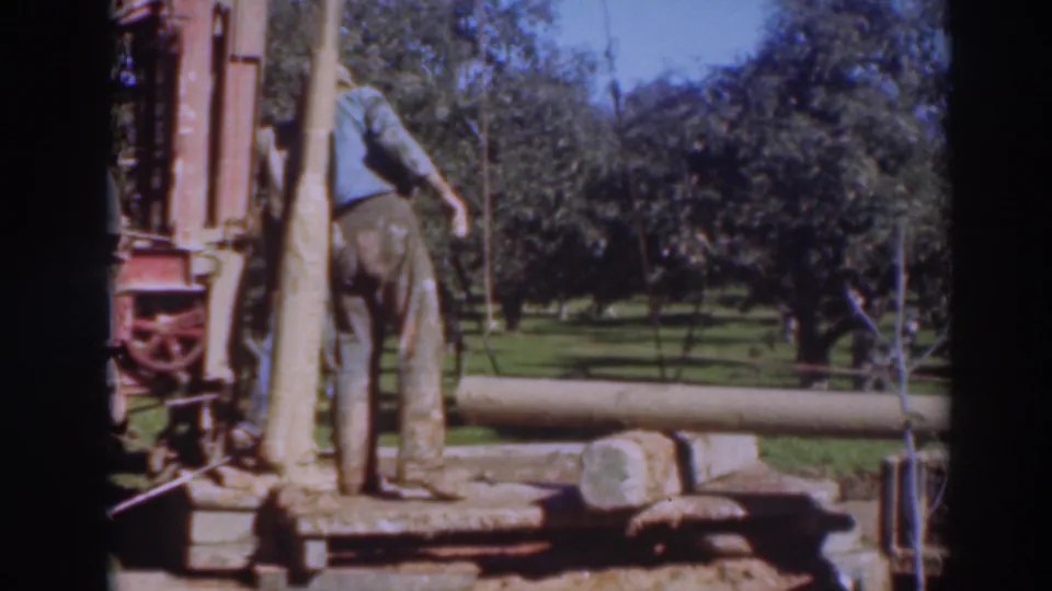 A man in work clothes stands by the portable saw rig and log in the 1937 Sacramento orchard scene, with trees filling the background.