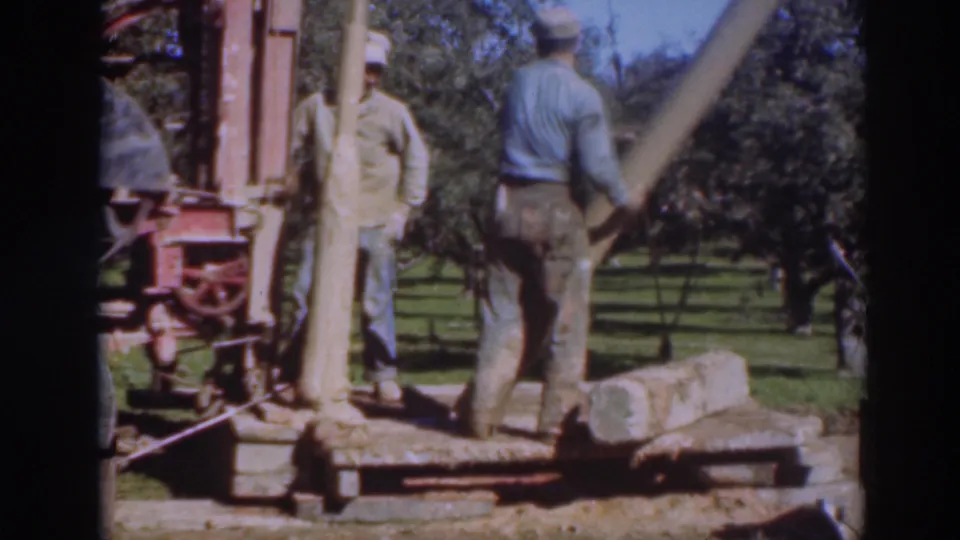 Two men in work clothes stand at the portable saw setup in a Sacramento orchard, with a log, cut timber, and rows of trees visible.