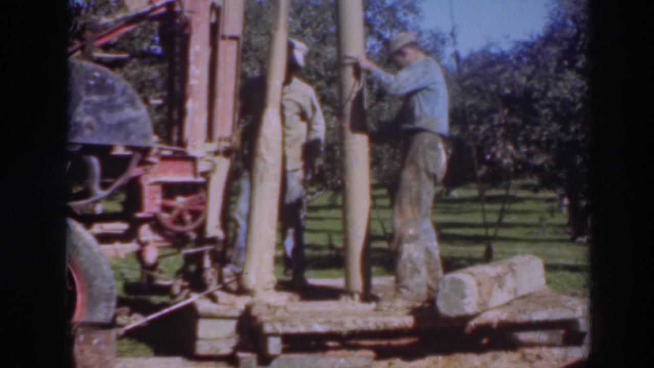 Two men stand beside upright wooden posts and a work rig in an orchard setting.