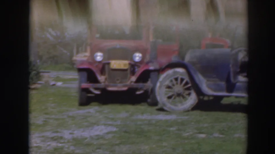 A red vintage vehicle and an older car sit on grass in the Sacramento orchard scene, partly blurred by film motion.