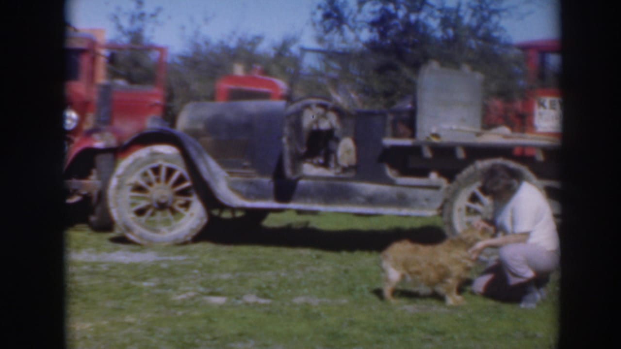 In 1937 Sacramento home-movie footage, a woman kneels to pet a small dog near a black automobile and red truck.
