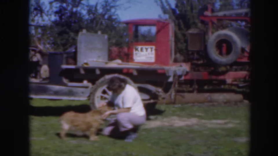 In the Sacramento yard scene, the woman bends toward the dog while the red truck, sign, and vehicle wheel are visible behind her.