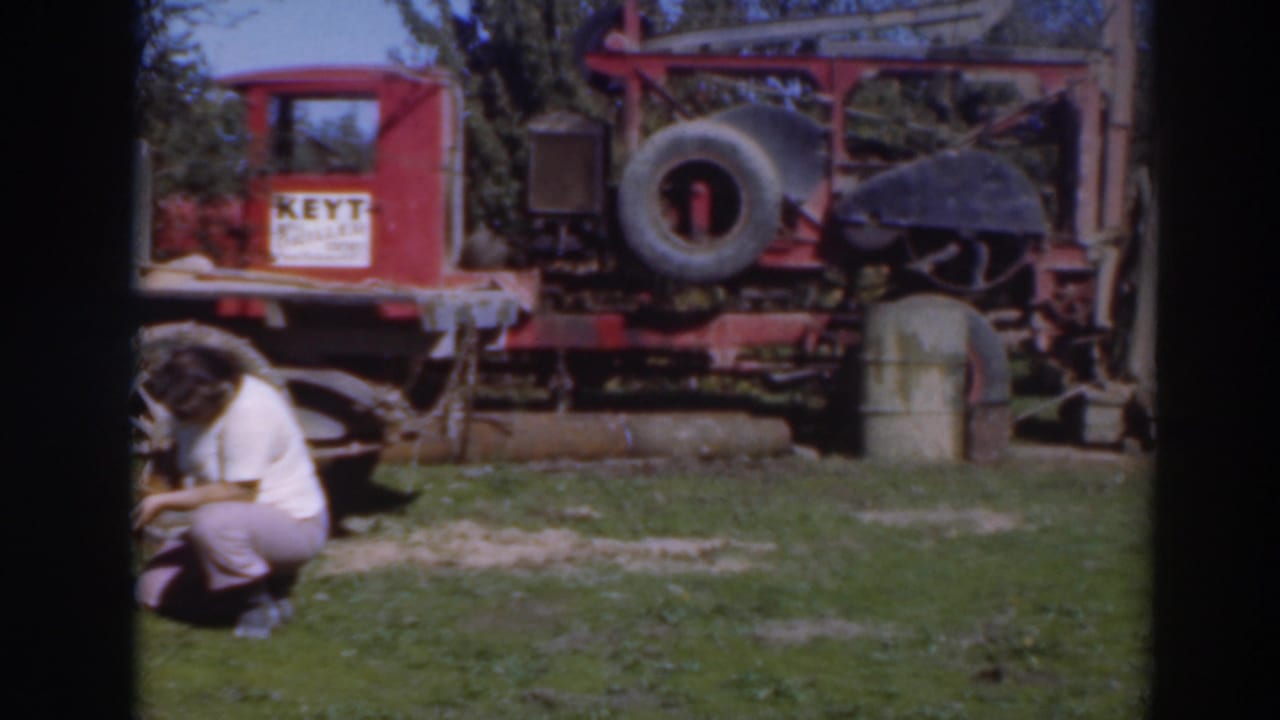 In 1937 Sacramento home-movie footage, a worker crouches near a red industrial rig with a visible sign, spare tire, barrel, and trees behind it.