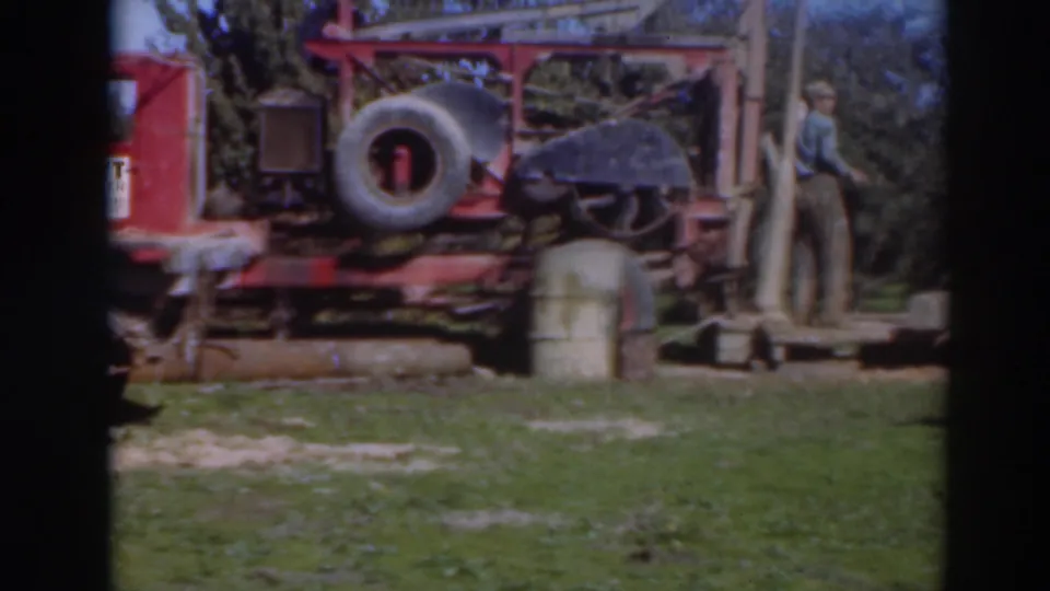 A man stands near the right side of the red industrial rig in the Sacramento 1937 footage, with a spare tire, barrel, trees, and grass visible.