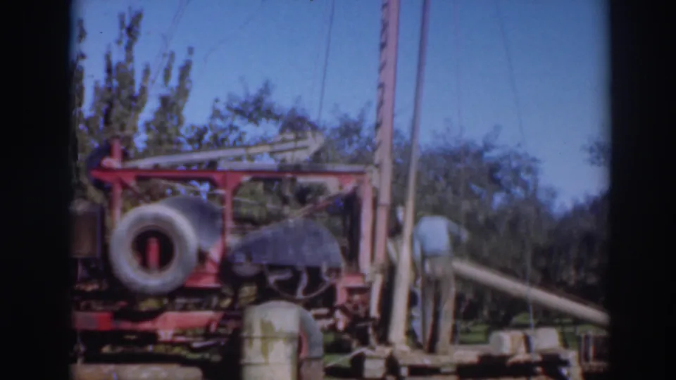 A worker bends near a long pipe or beam beside the red rig in 1937 Sacramento footage, with the spare tire, upright frame, grass, trees, and sky visible.
