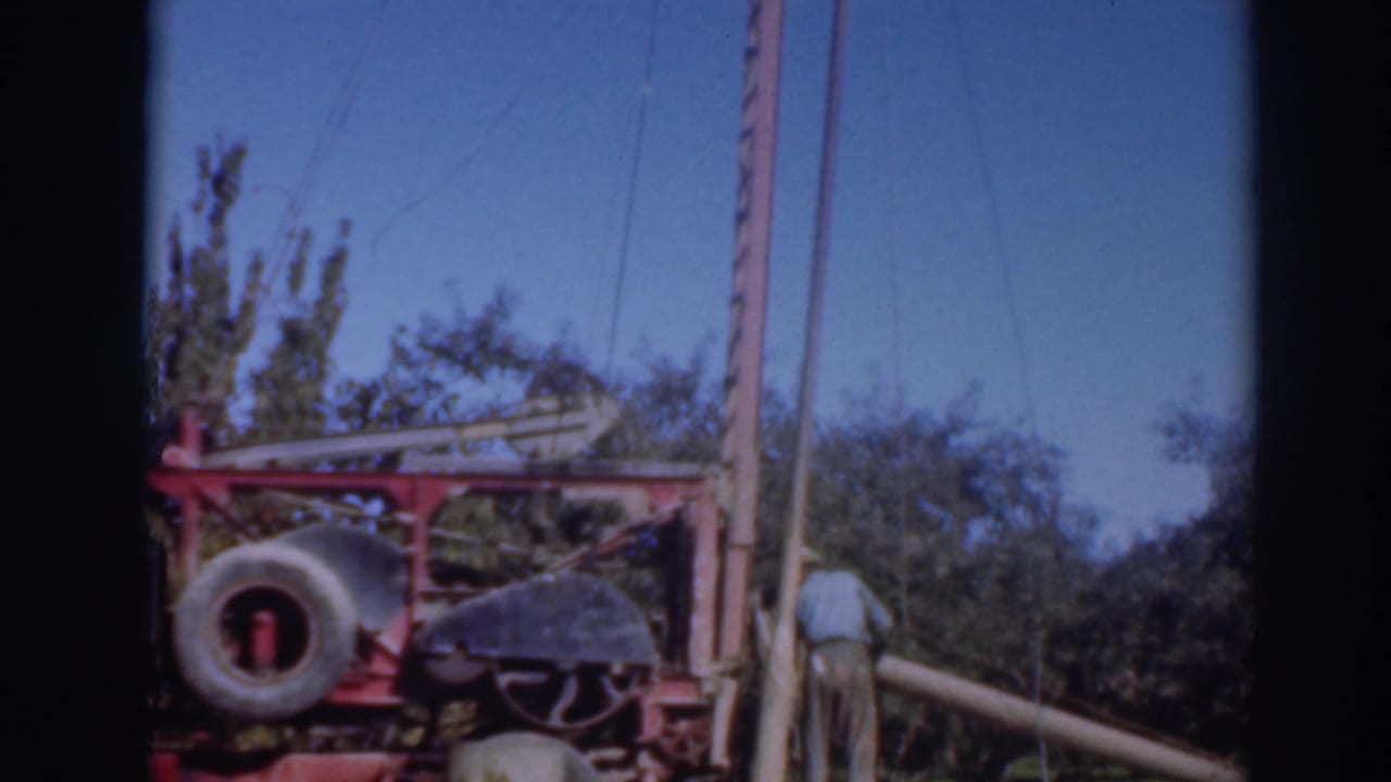 1937 Sacramento-area 8mm frame showing a worker near red outdoor machinery, a large tire, trees, poles, and clear sky.