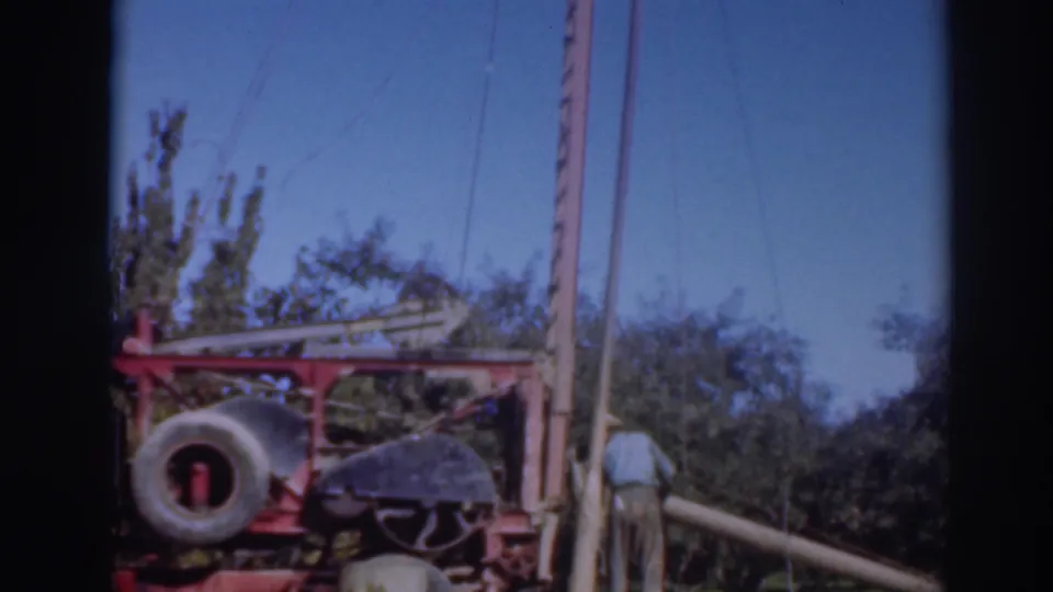 1937 Sacramento-area 8mm frame showing a worker near red outdoor machinery, a large tire, trees, poles, and clear sky.