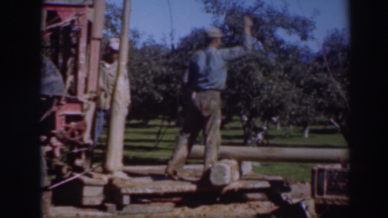 Man beside orchard machinery with another worker partly visible near trees.