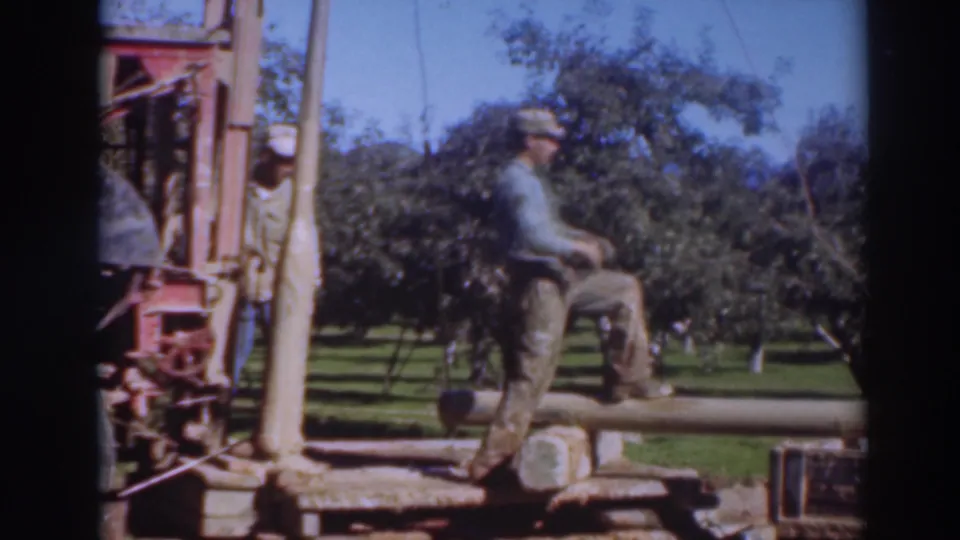 A worker in a cap and blue shirt steps on or over a horizontal timber near machinery in the Sacramento orchard scene.
