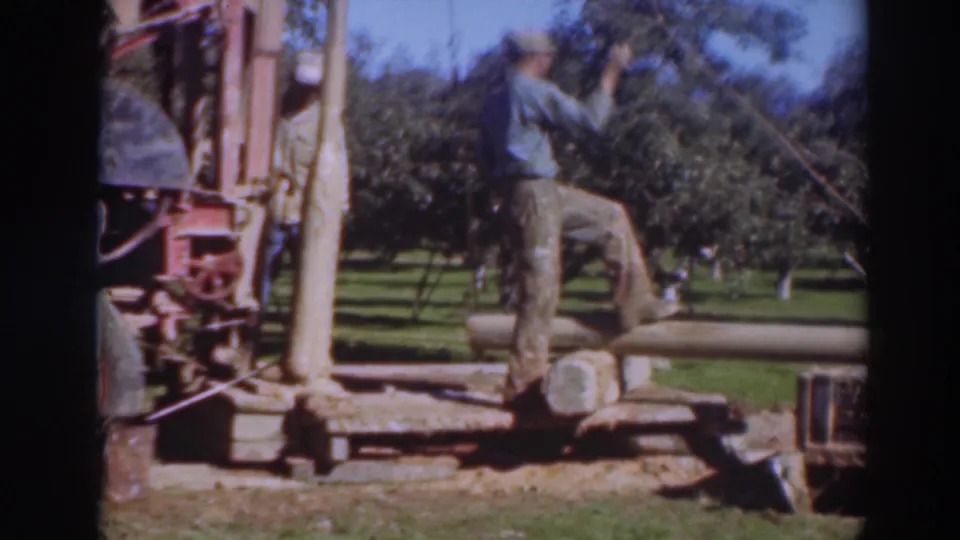 The worker appears to handle a cord or line beside the mechanical apparatus, with orchard rows visible behind him.