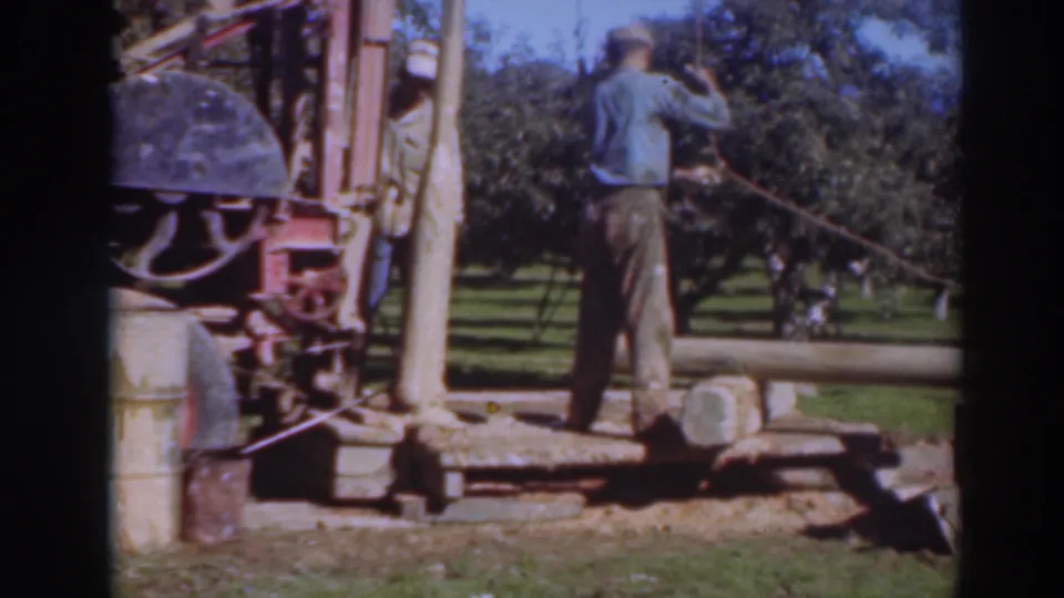 A man faces the equipment at the edge of an orchard while another worker is partly hidden near the machine.