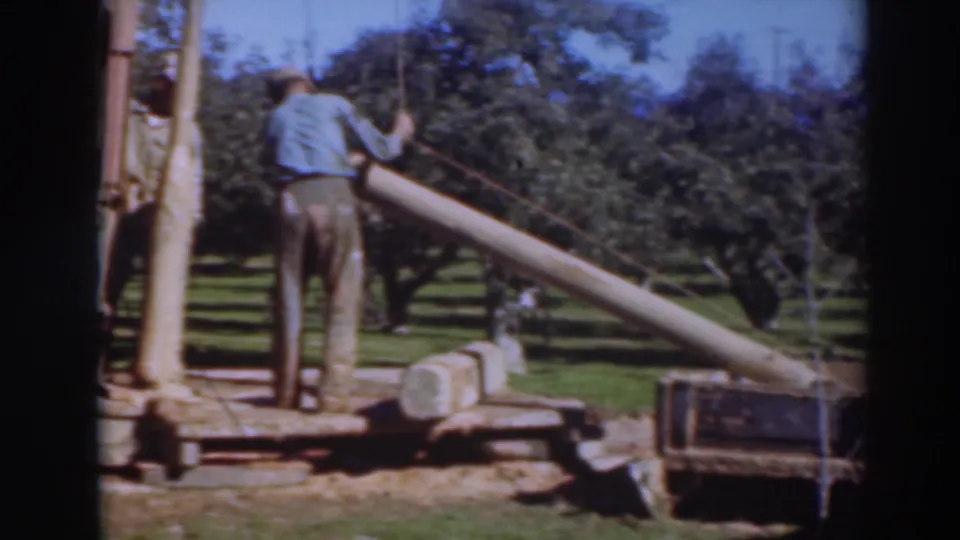 In the Sacramento orchard scene, the man stands beside the rig and lifts a long log upward toward the equipment platform.