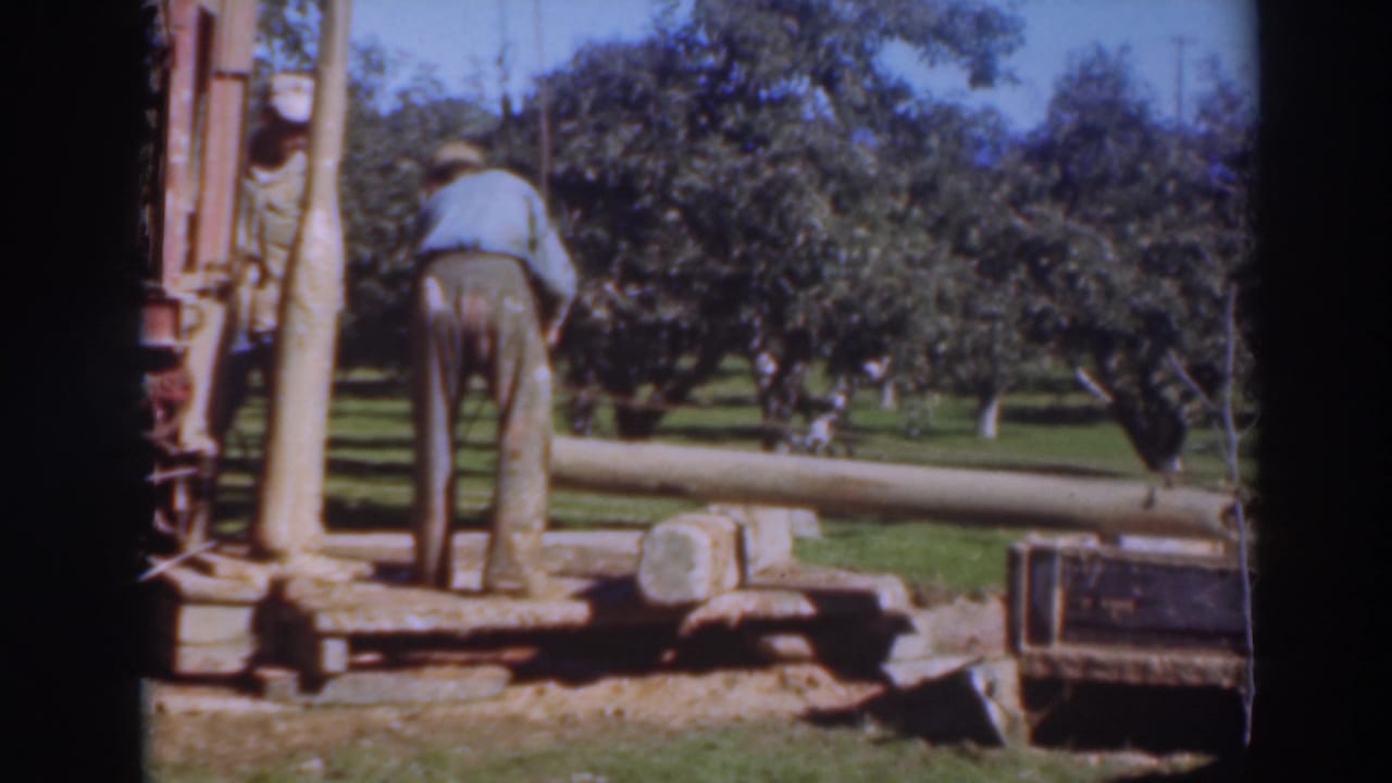 In 1937 Sacramento footage, a worker in a light shirt stands near a log loader and wooden beam in an orchard.