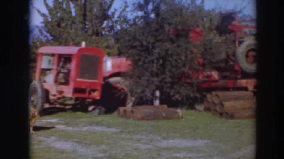 1937 Sacramento yard scene with a red tractor, red machinery, stacked logs, and trees in the background.