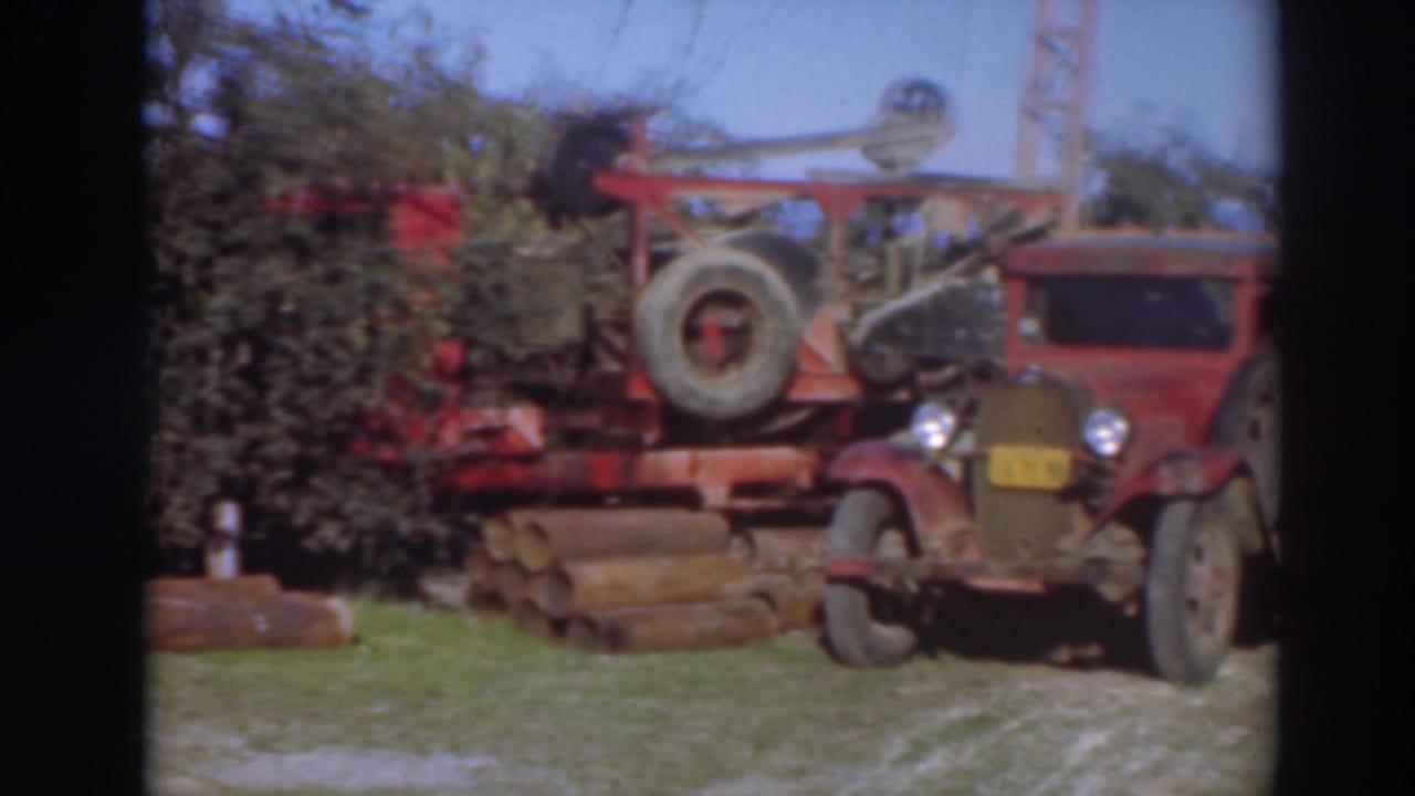 Red vintage truck parked beside logging machinery and stacked logs outdoors.