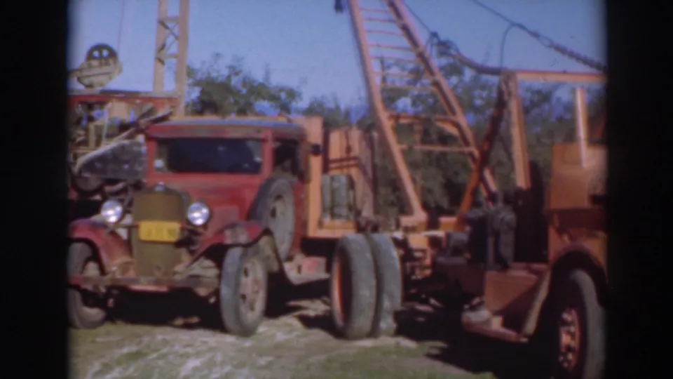1937 Sacramento frame with older trucks, tall rig-like equipment, trees, grass, and dark 8mm frame edges in a sunny outdoor setting.