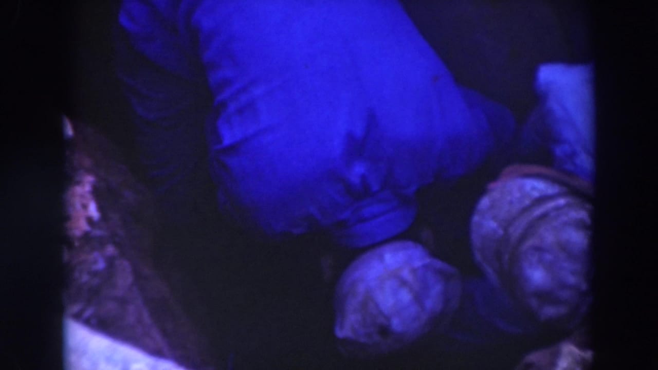 Workers in pale hats bend over in a dark pit-like area under blue-shifted light.