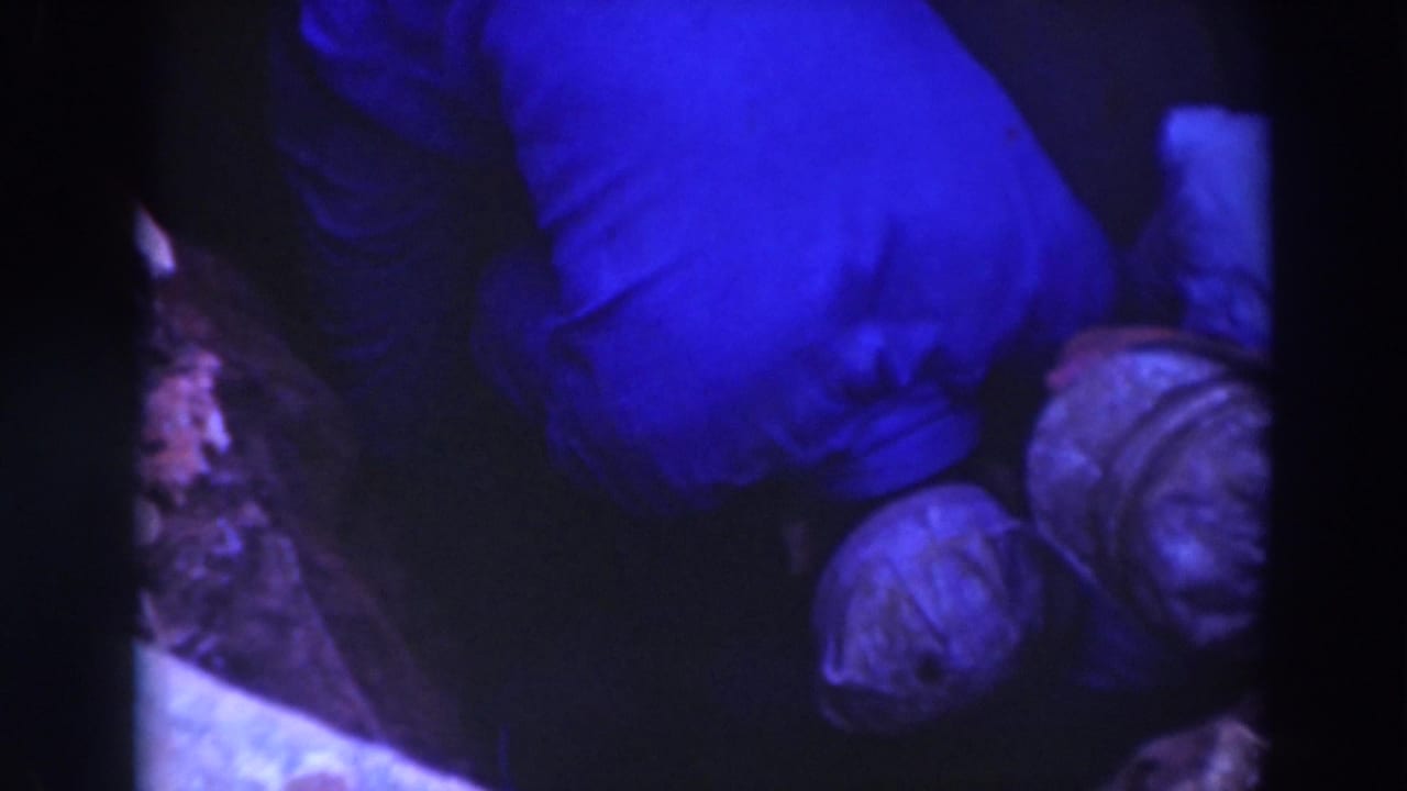 Two men in caps work in a dark, tight space with blue-tinted film color.