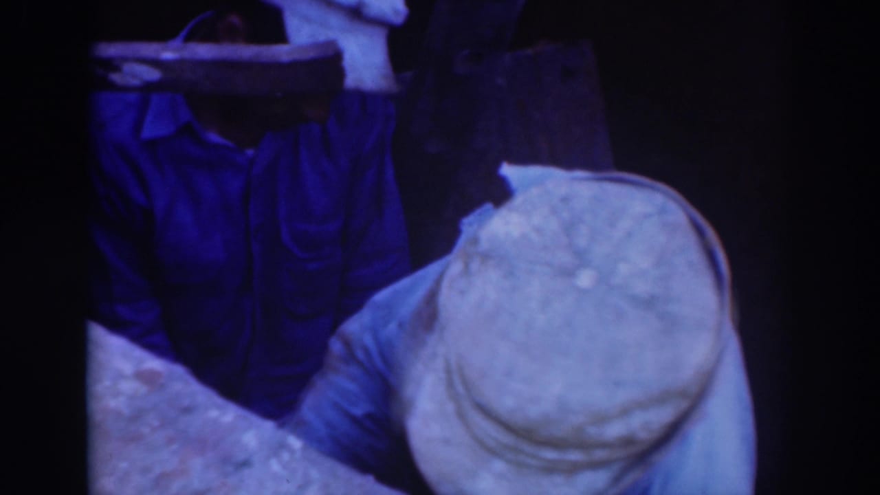 1937 Sacramento 8mm frame showing two capped workers in blue work clothing near dark machinery in a dim interior.