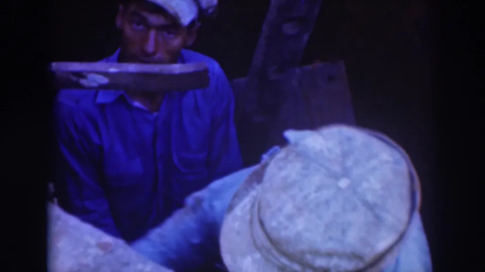 A capped worker in a blue uniform stands behind machinery while another capped worker bends over a task in the foreground.