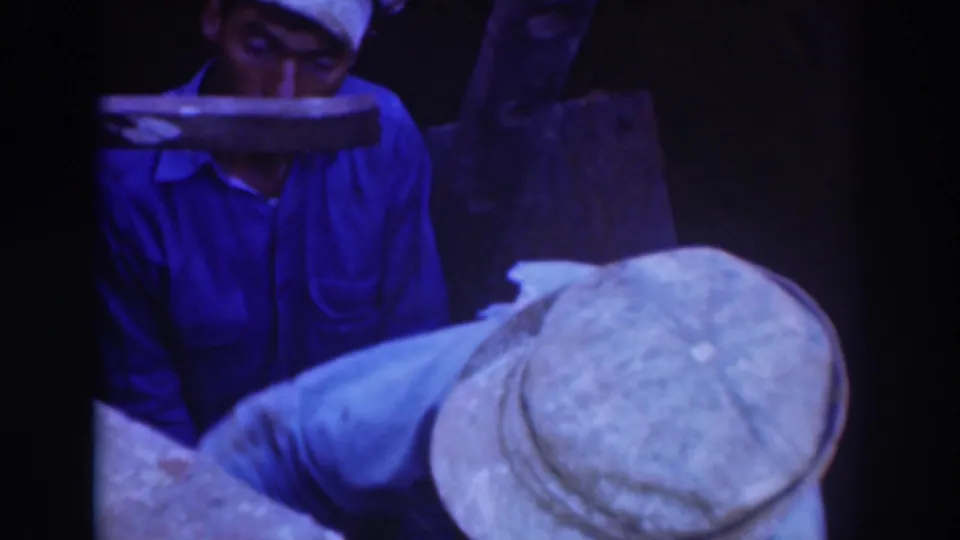 Dim 8mm frame with a blue-clad worker partly hidden by machinery and a second capped worker bent over the foreground task.