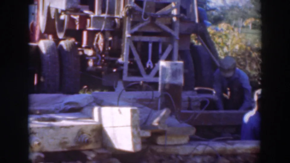 1937 Sacramento 8mm frame with men in work clothes crouched near machinery, cables, concrete blocks, and dirt ground.