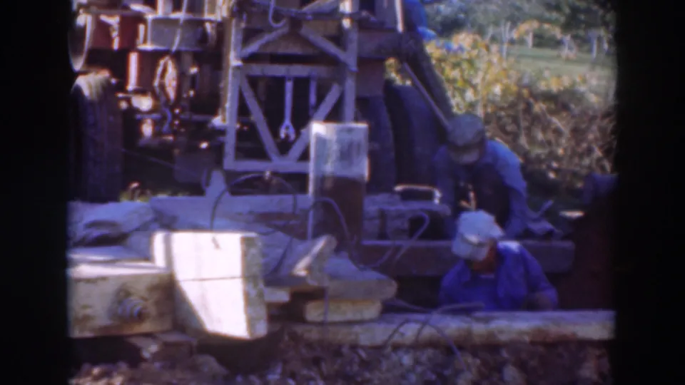 1937 Sacramento worksite frame showing workers beside a large rig, concrete forms, cables, and sunlit outdoor materials.