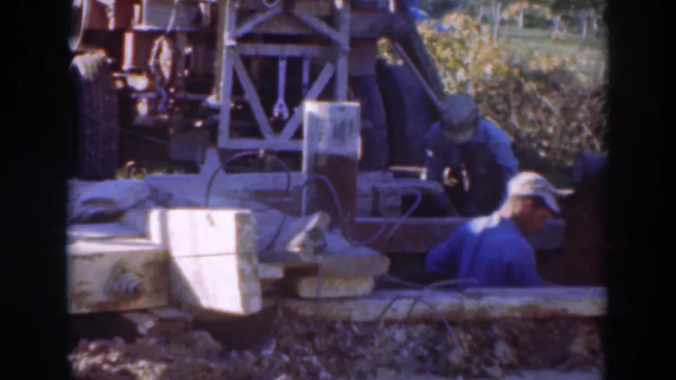 A worker in blue moves across the foreground at a Sacramento construction site, with drilling machinery and trench materials behind him.
