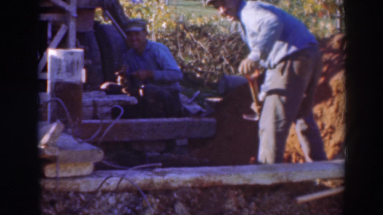 Two men work beside a wooden structure, one seated near a tool and another bending near exposed dirt.