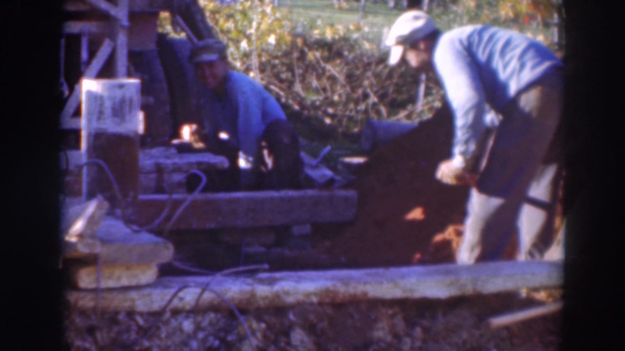 A man bends with a shovel near a dirt pile while another man sits by a wooden structure.