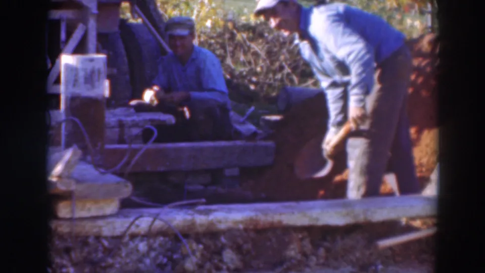 The foreground worker bends over a shovel as the seated worker watches or handles something beside the wooden structure.