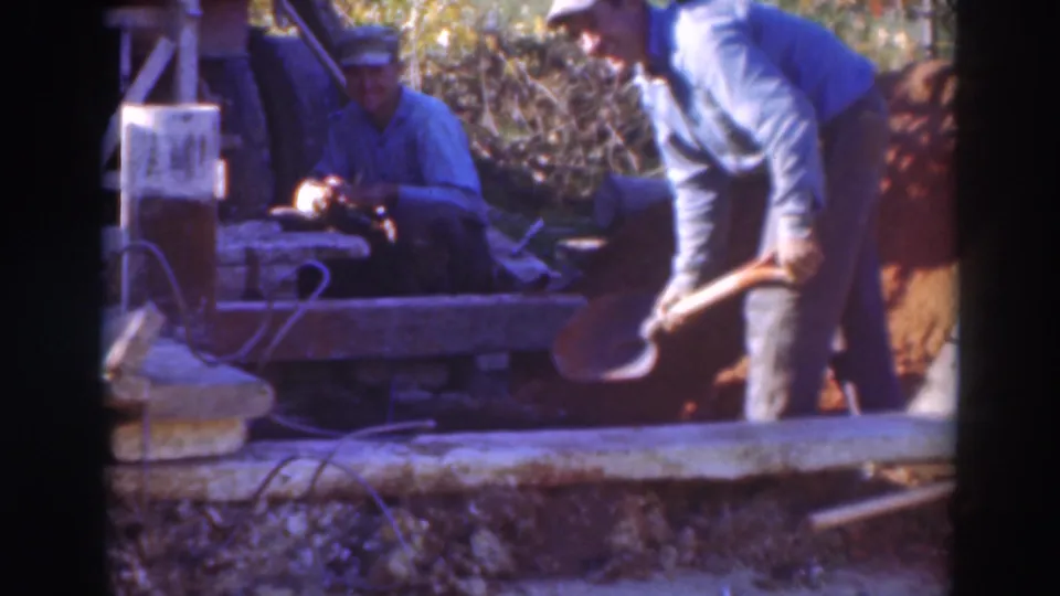 Near the end of the clip, the shoveling man stands close to the dirt bank while the seated man remains by the tool area.