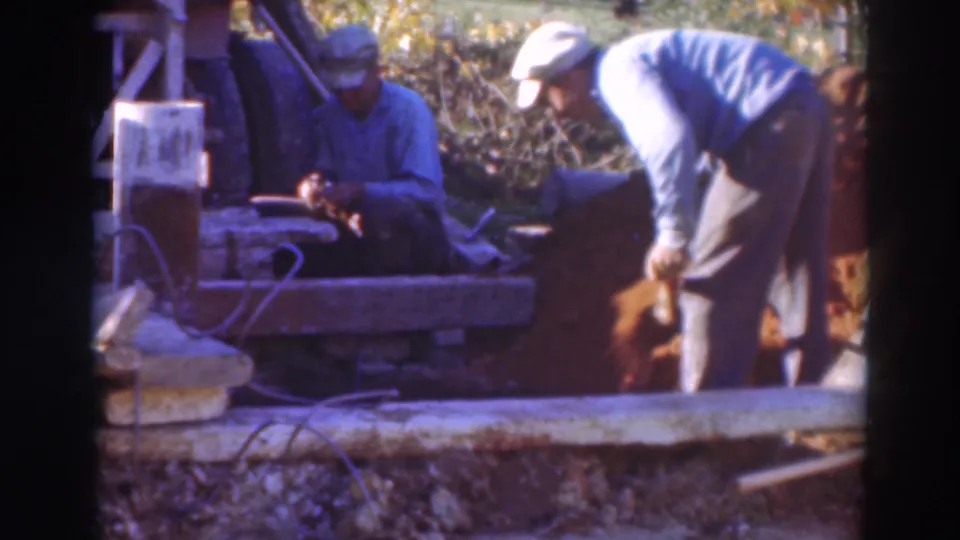 1937 Sacramento worksite view with one man seated near a wooden beam and another man in a light cap leaning over a shovel in loose earth.