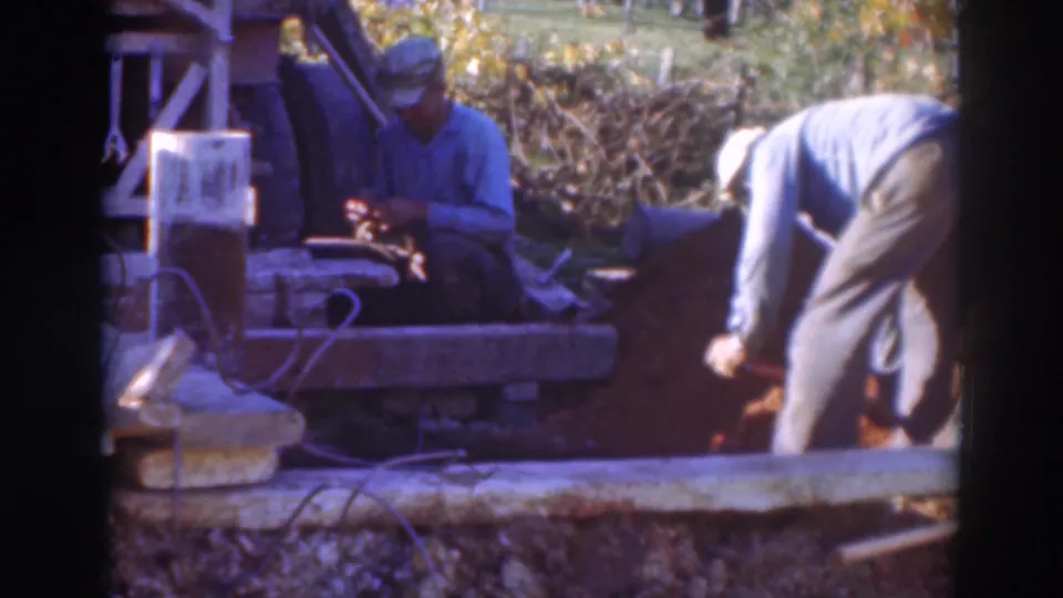 A 1937 Sacramento construction frame showing a worker bent forward over soil with a shovel and a seated worker beside timber and equipment.