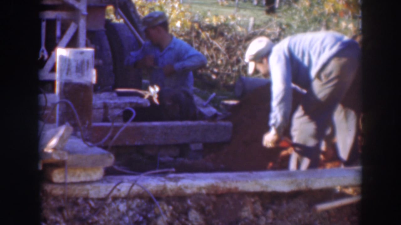 In 1937 Sacramento home-movie footage, one man sits by a worksite tool while another bends over loose soil with a shovel.