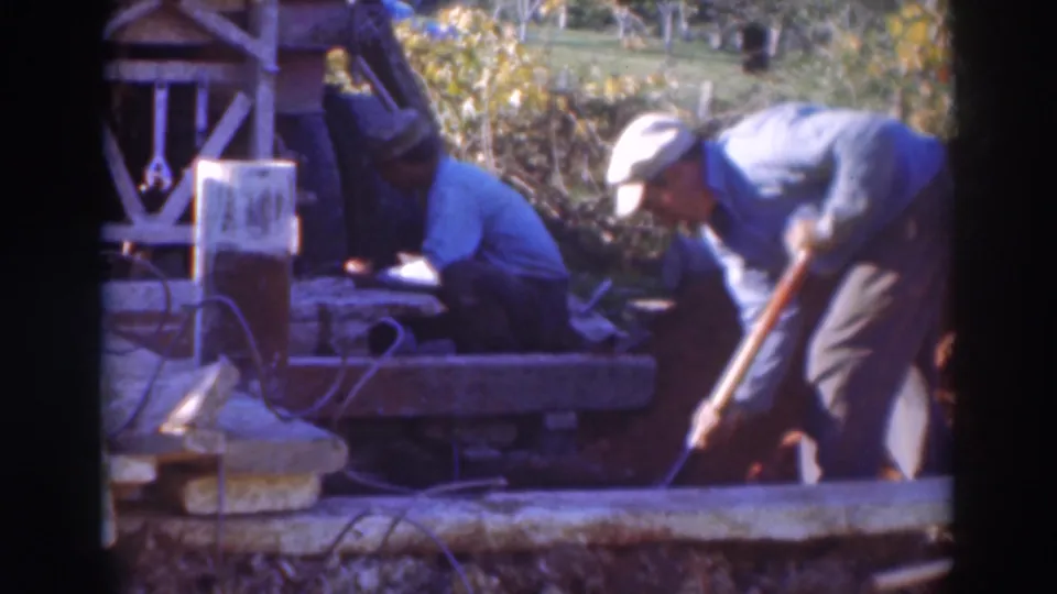 A man uses a long-handled tool in an outdoor Sacramento work area, with another man seated beside the wooden platform.