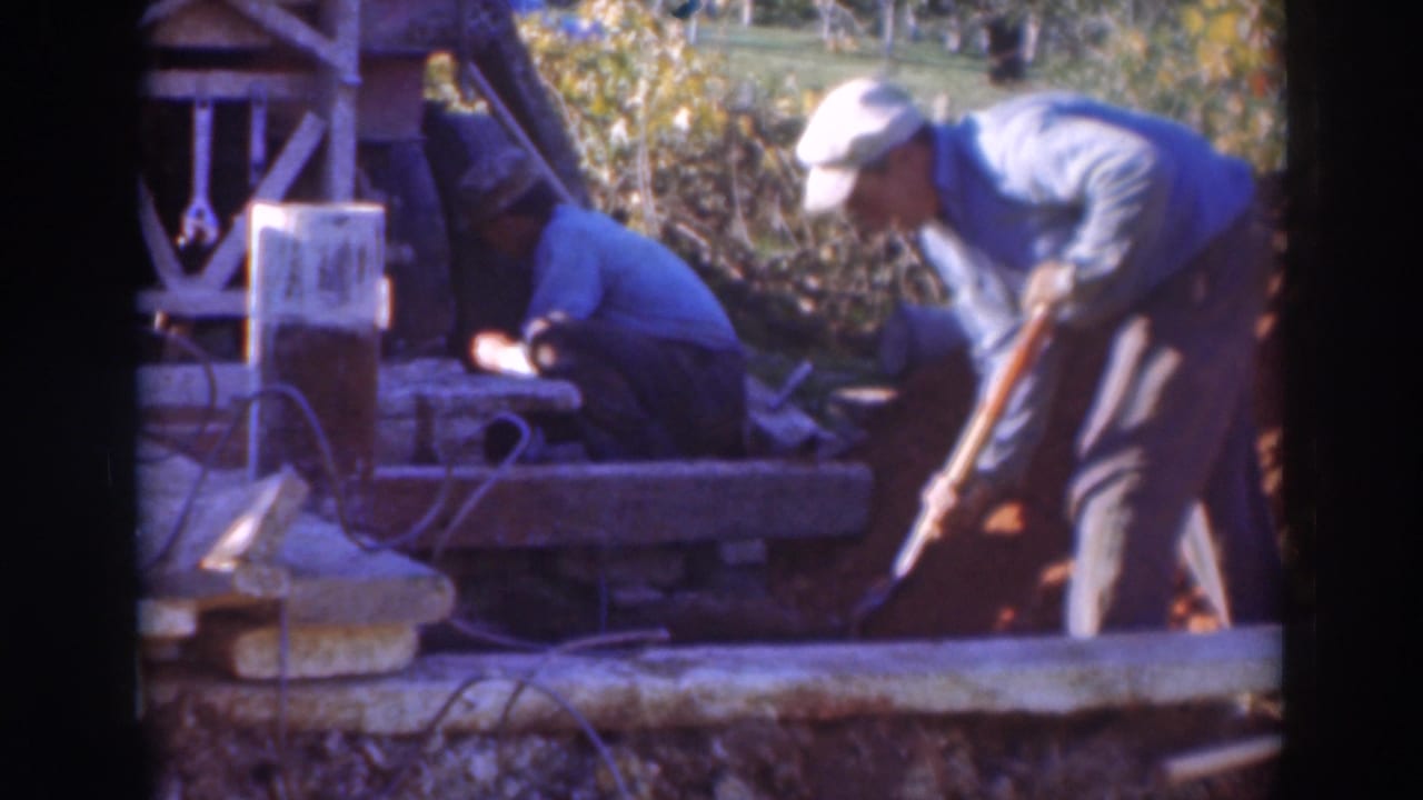 Two men work at an outdoor construction site, one kneeling near equipment while another uses a shovel.
