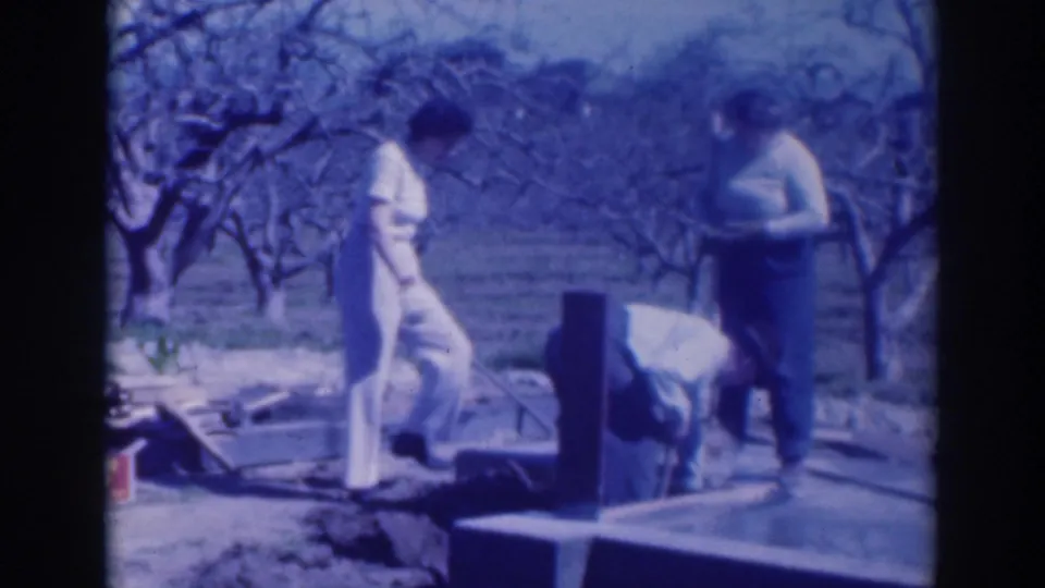 A wider 1937 Sacramento home-movie view shows two adults near construction materials, with rows of trees behind them.