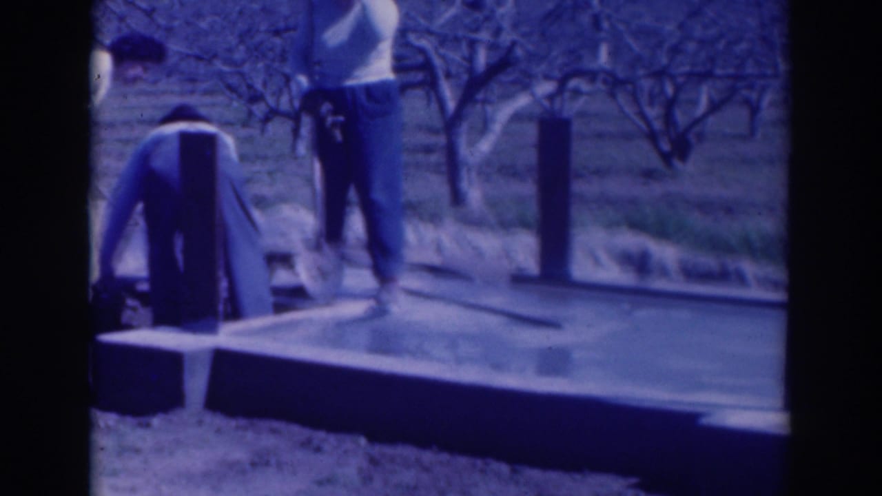 Two men work beside a wet concrete slab in an orchard, with a shovel and upright posts visible.