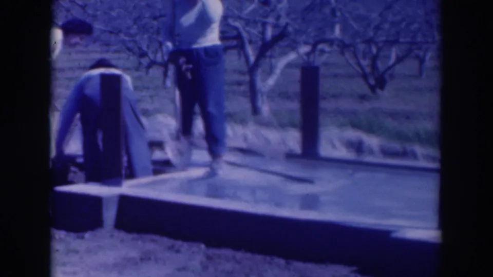 1937 Sacramento home-movie frame showing two men beside a freshly poured concrete foundation among bare orchard trees.