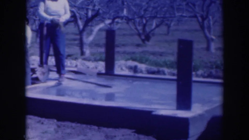 Blue-tinted 8mm frame from Sacramento showing a man at the edge of a concrete foundation with orchard trees beyond.