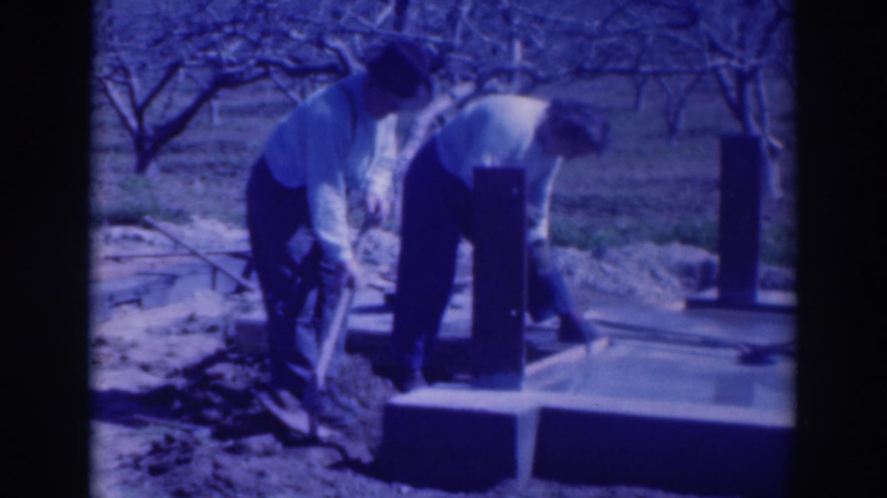 Two men bend over a concrete foundation in an orchard while using hand tools.