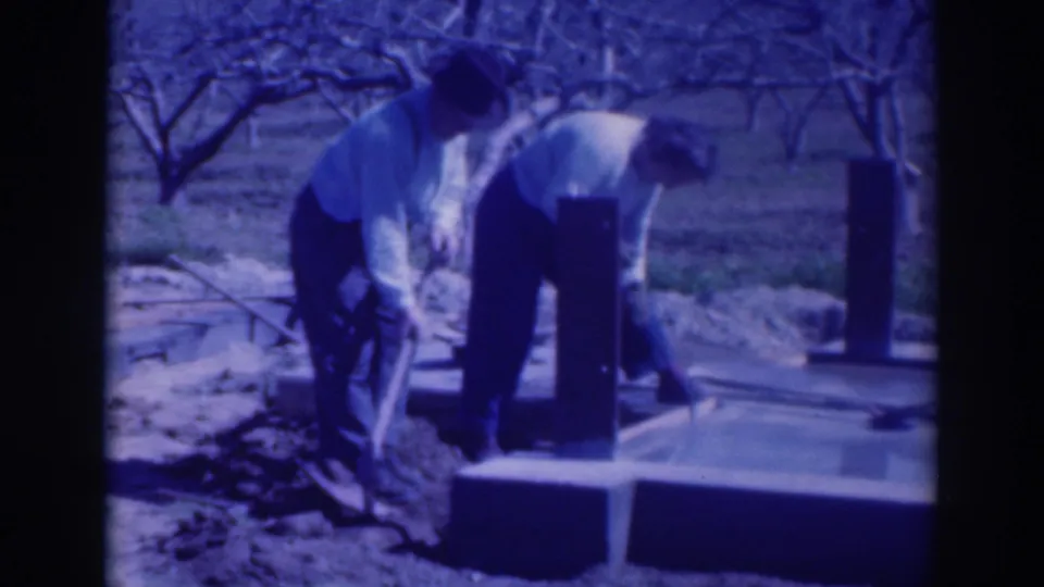Two adult men work with shovels beside a small concrete foundation in a Sacramento orchard, filmed on blue-tinted 8mm home movie footage.