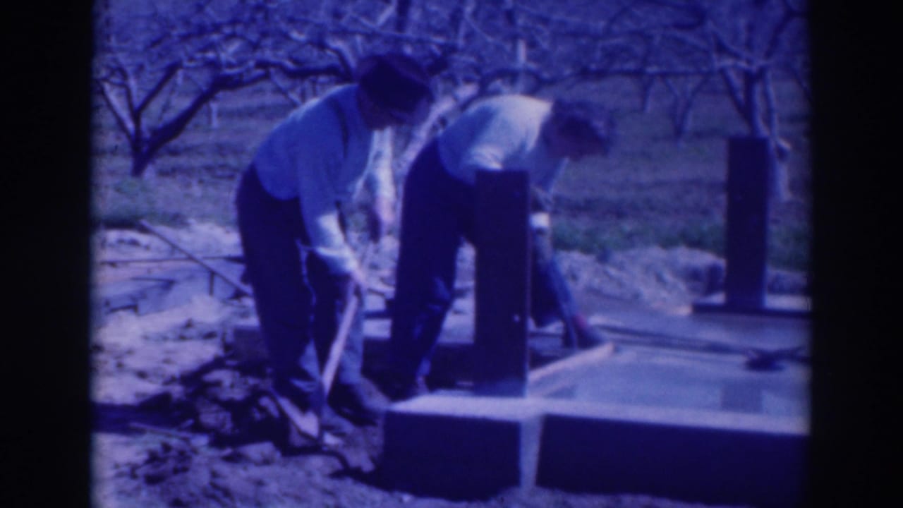 In the orchard setting, two men lean over the edge of a concrete slab or footing while working with shovels and other tools.