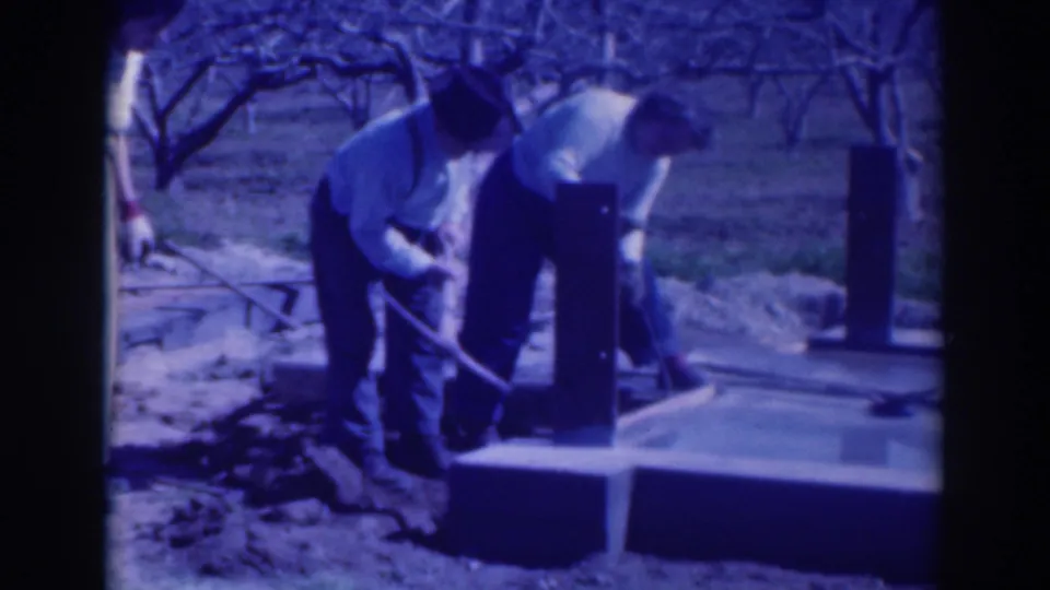 Two adult men in light shirts and dark trousers work near a concrete foundation, with bare orchard trees visible behind them.