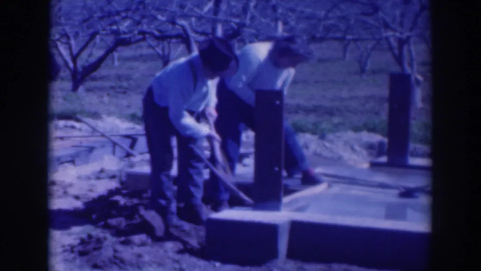 One man holds a shovel beside the concrete footing while the second man bends over the work area in the Sacramento orchard scene.