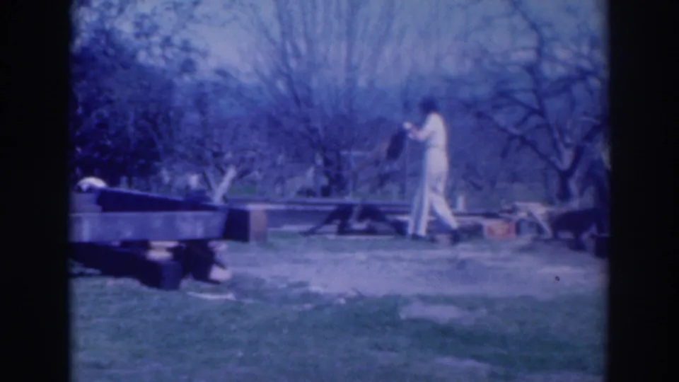 1937 Sacramento frame with a person in white pants and a light shirt handling debris beside a low wooden platform in a grassy yard.