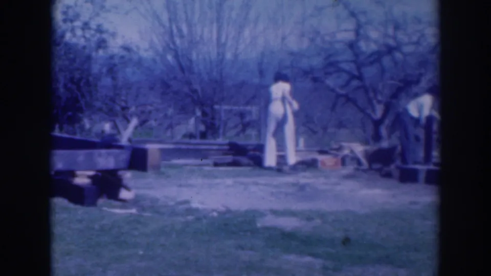 1937 Sacramento home-movie frame showing two adults in a grassy backyard near scattered boards or blocks under bare trees.