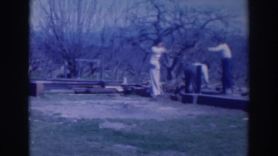 Home-movie frame from Sacramento in 1937 showing adults handling wood pieces beside grass, bare trees, and a low dark edge.